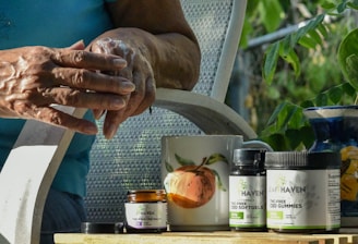 A person is applying cream to their hands while seated outside. A small jar of cream, along with bottles labeled CBD softgels and gummies, are on a table. A mug with a peach design and a ceramic vase are also present on the table. Green foliage is visible in the background, suggesting a garden setting.