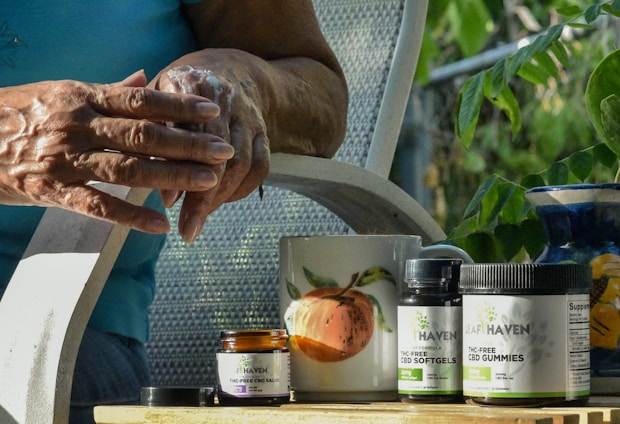 A person is applying cream to their hands while seated outside. A small jar of cream, along with bottles labeled CBD softgels and gummies, are on a table. A mug with a peach design and a ceramic vase are also present on the table. Green foliage is visible in the background, suggesting a garden setting.