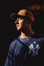 A person is illuminated by a spotlight, wearing a brown cap and a navy blue sports jersey with a prominent logo. The background is dark, which emphasizes the contrast with the lighter foreground.