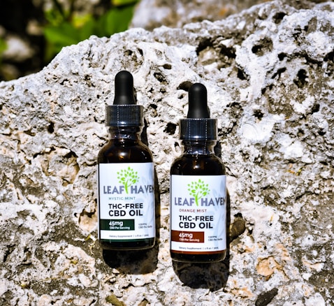 Two bottles of Leaf Haven THC-Free CBD Oil are placed on a textured rock surface. Each bottle has a dropper cap and distinct labeling. The bottle on the left is labeled Mystic Mint, while the one on the right is labeled Orange Mist. The background is a rough, uneven stone, adding a natural and earthy feel to the setting.