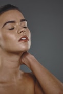 A person with closed eyes appears to be in a serene or contemplative state, with their head tilted slightly and hand resting on the neck. The skin has a smooth and glowing appearance.