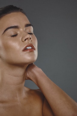 A person with closed eyes appears to be in a serene or contemplative state, with their head tilted slightly and hand resting on the neck. The skin has a smooth and glowing appearance.