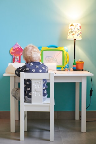 A young child with blonde hair sits at a small white table, engaged in drawing or writing on paper. The child is facing away, wearing a dark blue shirt with a cartoon eyes pattern. The table has various items on it, including a colorful magnetic drawing board, a small lamp with a floral design, and some colorful toys or markers. The background wall is painted a bright turquoise color, adding a vibrant backdrop to the scene.