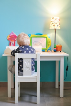 A young child with blonde hair sits at a small white table, engaged in drawing or writing on paper. The child is facing away, wearing a dark blue shirt with a cartoon eyes pattern. The table has various items on it, including a colorful magnetic drawing board, a small lamp with a floral design, and some colorful toys or markers. The background wall is painted a bright turquoise color, adding a vibrant backdrop to the scene.