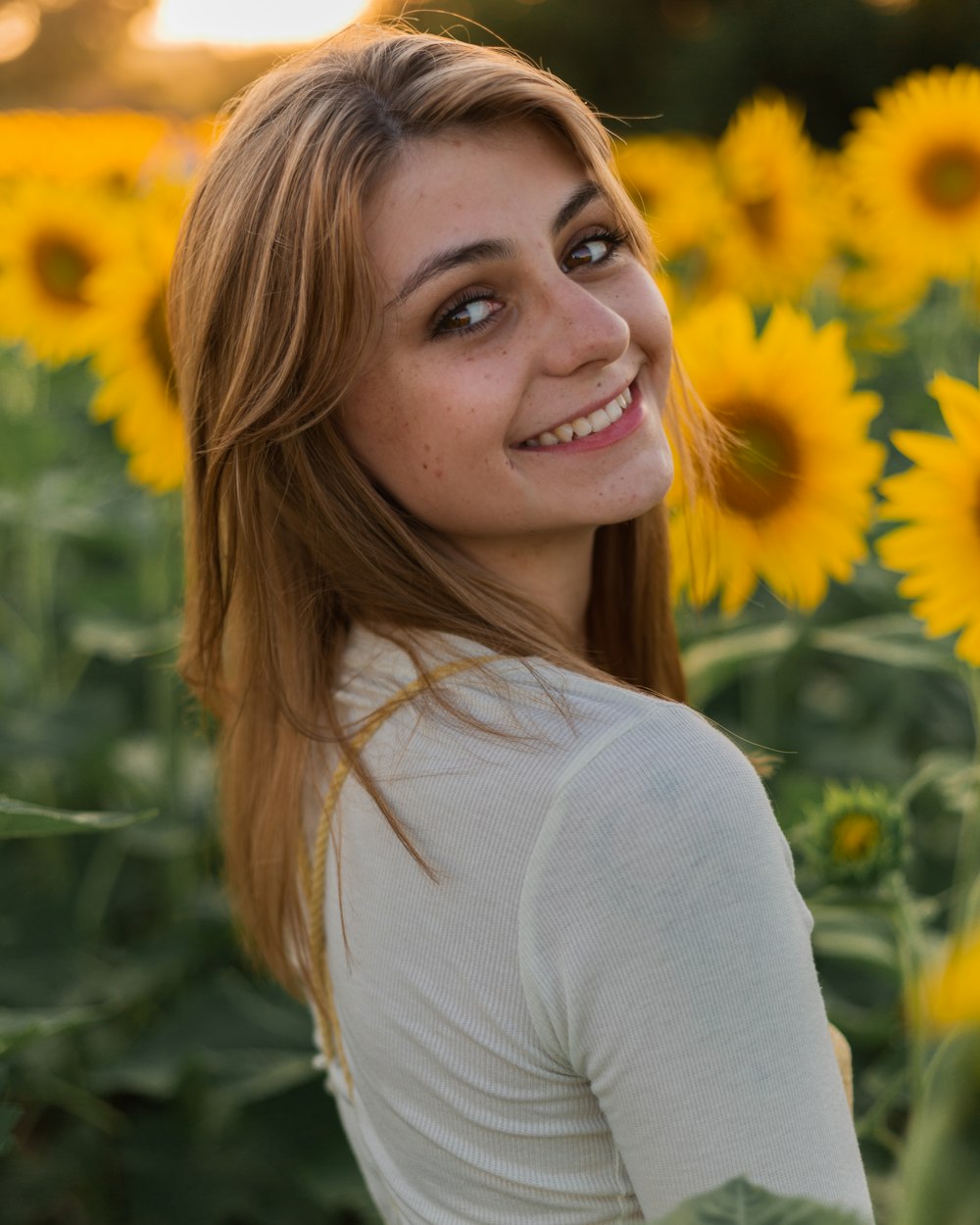昼間、黄色い花のそばに立つ白い長袖シャツを着た笑顔の女性
