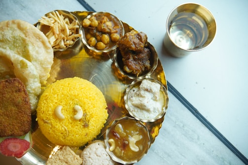 A traditional Indian meal is served on a metal platter, featuring various dishes. The central focus is yellow rice garnished with cashews. Surrounding it are puris, a deep-fried bread, a variety of curries with chickpeas and vegetables, potato fries, and sweets, each in separate compartments. A shiny brass cup containing water is placed beside the platter on a marble surface.