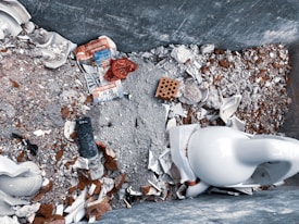 Construction debris fills a large container, including broken ceramic pieces, a discarded newspaper, and a pipe. A brick and various rubble materials add to the chaotic mix.