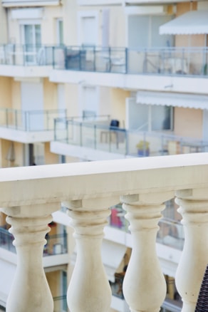 A close-up view of a white balcony railing with detailed balustrades, overlooking an apartment building with multiple balconies. The building has a light beige facade with glass railings and some balconies have furniture like chairs and tables.