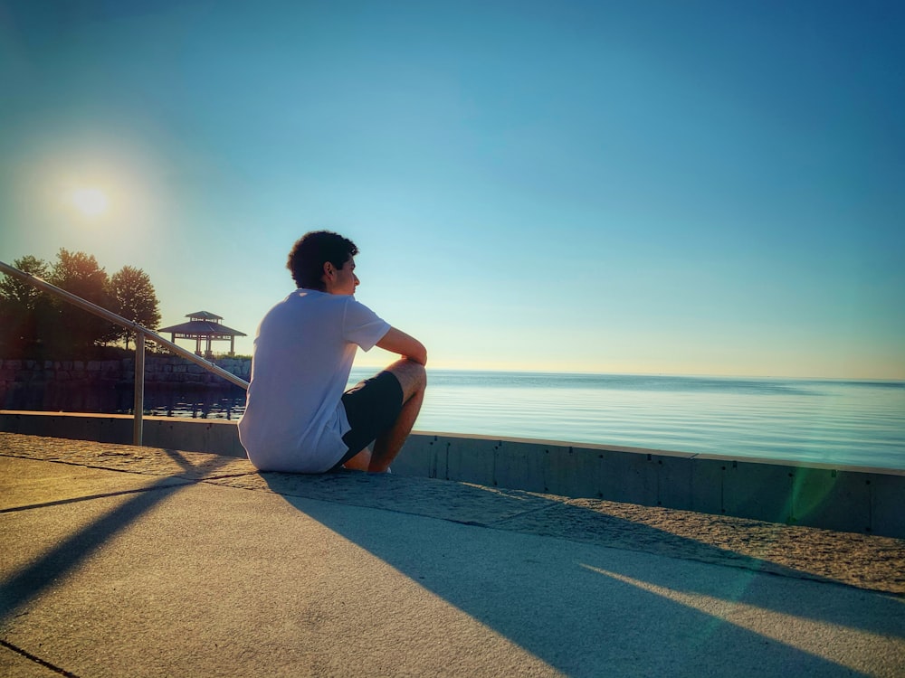 man in white t-shirt sitting on concrete pavement near body of water during daytime