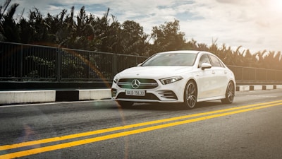 A sleek white sedan is driving on a road with double yellow lines, bordered by a metal guardrail. The sky appears cloudy with sunlight filtering through, and the surrounding area is lined with lush greenery.