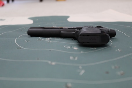 A handgun is placed on a shooting target with visible bullet holes and concentric circles. The focus is on the firearm lying flat on the target surface.