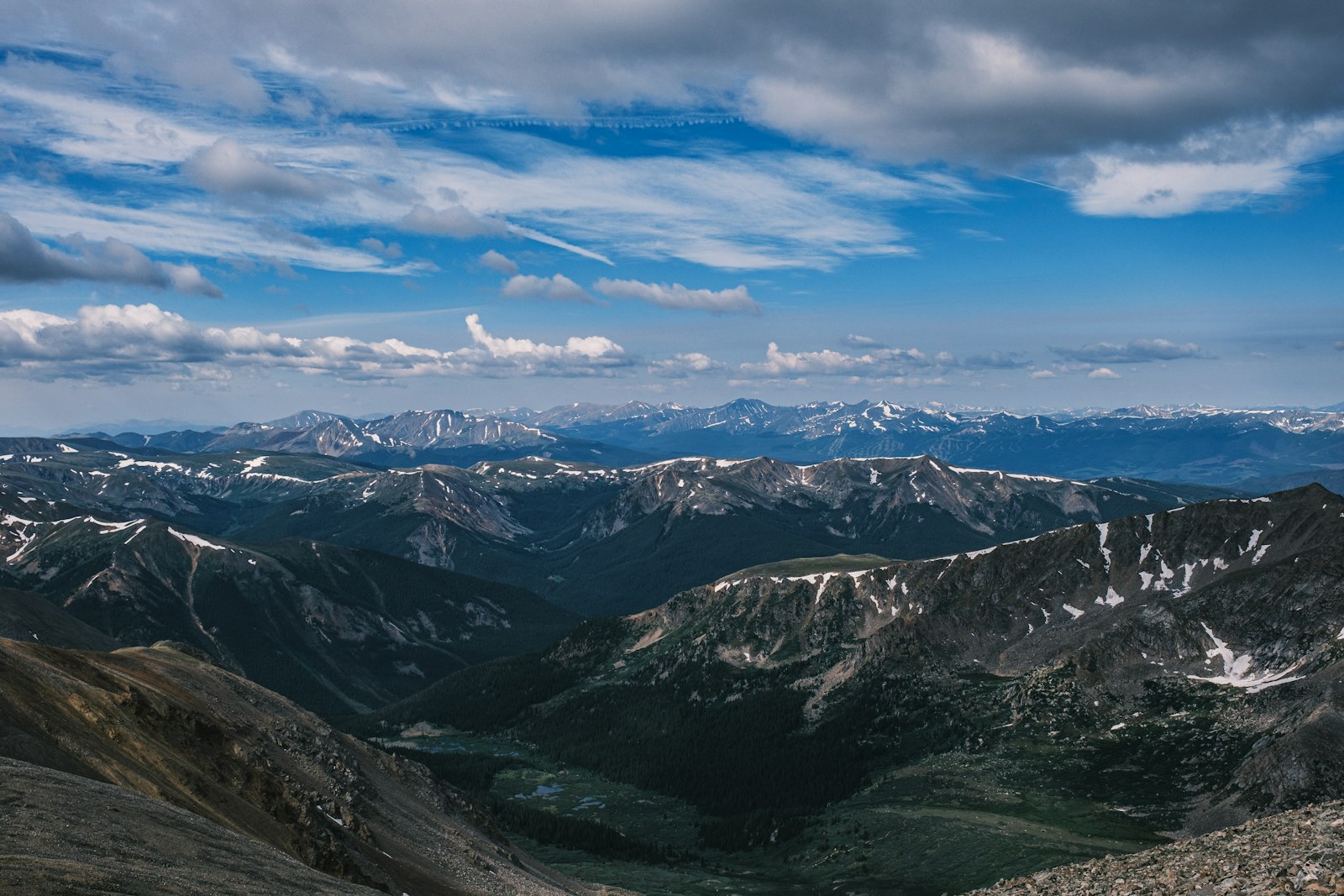 A sweeping high-altitude panorama of layered mountain ridges and green valleys with lingering snow patches
