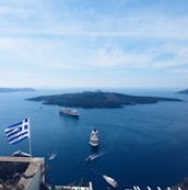 A picturesque view of a Greek island in the Aegean Sea, featuring several large cruise ships sailing near the coast. The foreground shows a Greek flag fluttering in the breeze, indicating a viewpoint from a higher elevation. The calm blue waters contrast with the rugged outline of the island under a clear and bright sky.