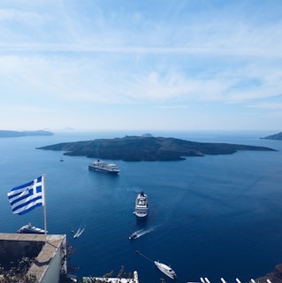 A picturesque view of a Greek island in the Aegean Sea, featuring several large cruise ships sailing near the coast. The foreground shows a Greek flag fluttering in the breeze, indicating a viewpoint from a higher elevation. The calm blue waters contrast with the rugged outline of the island under a clear and bright sky.