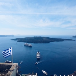 A picturesque view of a Greek island in the Aegean Sea, featuring several large cruise ships sailing near the coast. The foreground shows a Greek flag fluttering in the breeze, indicating a viewpoint from a higher elevation. The calm blue waters contrast with the rugged outline of the island under a clear and bright sky.