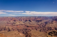 Vast and expansive landscape featuring the Grand Canyon with its layered bands of red, orange, and brown rock formations. The sky is clear with a few clouds scattered, offering a stark contrast to the rugged terrain below.