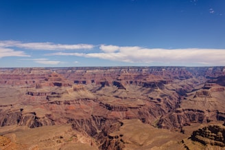 Vast and expansive landscape featuring the Grand Canyon with its layered bands of red, orange, and brown rock formations. The sky is clear with a few clouds scattered, offering a stark contrast to the rugged terrain below.