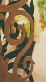 An intricately designed ornamental metal piece with swirling patterns, placed in front of a blurred background of greenery and flowers. The metal looks rusty with a brown patina.