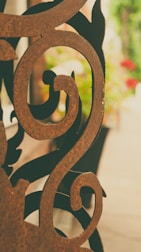 An intricately designed ornamental metal piece with swirling patterns, placed in front of a blurred background of greenery and flowers. The metal looks rusty with a brown patina.