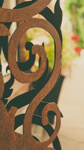 An intricately designed ornamental metal piece with swirling patterns, placed in front of a blurred background of greenery and flowers. The metal looks rusty with a brown patina.