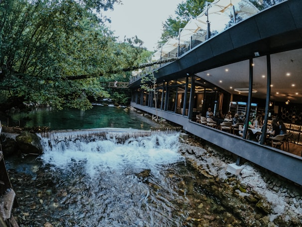 A modern restaurant with large glass windows overlooks a serene river with a small waterfall. People are dining inside, enjoying the view of nature that is rich with greenery and surrounded by trees.