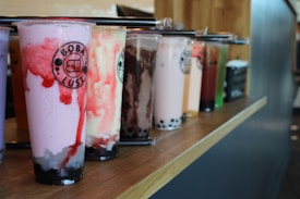 A row of assorted bubble tea cups sit on a wooden counter. Each cup contains different flavors and colors, with ingredients such as tapioca pearls visible at the bottom. The drinks are topped with plastic covers and have black straws wrapped in plastic.