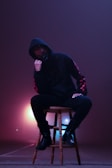 A person wearing a dark hoodie and black boots is sitting on a wooden stool. The background has an ethereal glow with hues of purple and pink, creating a misty atmosphere. The person's face is partially obscured, adding a mysterious vibe.