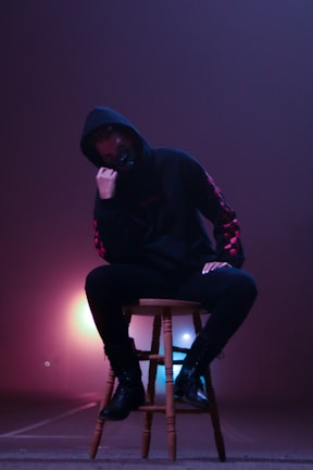 A person wearing a dark hoodie and black boots is sitting on a wooden stool. The background has an ethereal glow with hues of purple and pink, creating a misty atmosphere. The person's face is partially obscured, adding a mysterious vibe.
