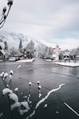 A snow-covered street in a small town with mountains in the background. Sparse traffic is visible, with parked cars along the roadside. The sky is cloudy, and bare trees line the street. A few storefronts and signs are visible, hinting at a quiet, serene atmosphere.