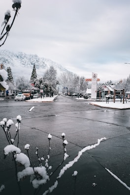 A snow-covered street in a small town with mountains in the background. Sparse traffic is visible, with parked cars along the roadside. The sky is cloudy, and bare trees line the street. A few storefronts and signs are visible, hinting at a quiet, serene atmosphere.