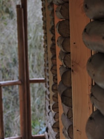 A close-up view of a wooden structure, built with logs stacked horizontally. The focus is on the texture and natural color of the wood, capturing the rustic craftsmanship. In the background, there is a blurred view of foliage, indicating an outdoor setting.