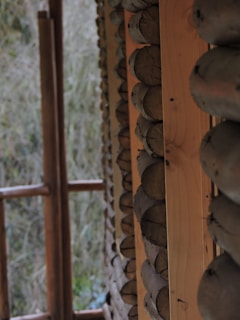 A close-up view of a wooden structure, built with logs stacked horizontally. The focus is on the texture and natural color of the wood, capturing the rustic craftsmanship. In the background, there is a blurred view of foliage, indicating an outdoor setting.
