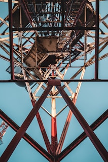 A low-angle view of a steel tower with a grid-like structure. The metal beams crisscross in geometric patterns, creating an intricate design. The backdrop is a clear blue sky, enhancing the metal framework's industrial appearance.