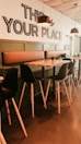 A cozy cafe interior with a modern design features high wooden tables and black chairs with wooden legs. Pendant lights hang from the ceiling, and a wall with a large, welcoming sign saying 'THIS IS YOUR PLACE' adds a personal touch.