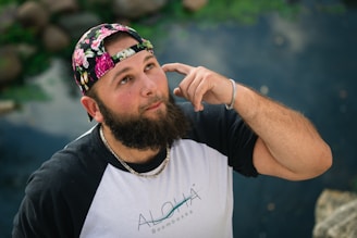 A man with a beard is gazing upwards while placing his index finger near his ear. He wears a floral-patterned cap and a black and white raglan sleeve shirt with a visible necklace. The background consists of a blurred natural environment with rocks and water.