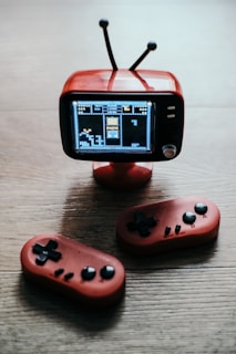 A small, vintage-style television with an antenna displays a pixelated game screen. Below are two red handheld gaming controllers on a wooden surface.