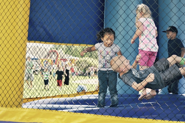 Several young children are playing inside a bouncy house. The bouncing space is enclosed with a yellow and blue net, and the children seem to be enjoying themselves. The background shows a busy outdoor area with more children and adults, hinting at a fair or a community event.