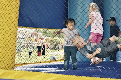 Several young children are playing inside a bouncy house. The bouncing space is enclosed with a yellow and blue net, and the children seem to be enjoying themselves. The background shows a busy outdoor area with more children and adults, hinting at a fair or a community event.