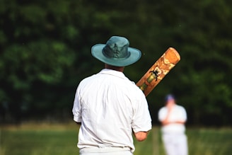 A person wearing a wide-brimmed hat and a white shirt stands with a cricket bat featuring colorful artwork on the back. The person is facing away, focusing on a distant figure dressed in similar attire, with a grassy field and blurred trees in the background.