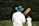 A person wearing a wide-brimmed hat and a white shirt stands with a cricket bat featuring colorful artwork on the back. The person is facing away, focusing on a distant figure dressed in similar attire, with a grassy field and blurred trees in the background.