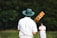 A person wearing a wide-brimmed hat and a white shirt stands with a cricket bat featuring colorful artwork on the back. The person is facing away, focusing on a distant figure dressed in similar attire, with a grassy field and blurred trees in the background.