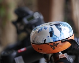 A bicycle bell is mounted on a handlebar. The bell has a distinct design, featuring a top half decorated with blue and white patterns, possibly including images of animals or nature. The bottom half is orange with a few scratches and paint marks. The background is blurred, suggesting an outdoor setting with some greenery.
