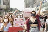 A group of people is gathered in a public area, holding various signs with messages advocating for women's rights and against assault. Many are wearing face masks, and there is a mix of emotions displayed, including determination and solidarity. The backdrop includes urban buildings with several people raising their fists in the air.