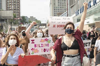 A group of people is gathered in a public area, holding various signs with messages advocating for women's rights and against assault. Many are wearing face masks, and there is a mix of emotions displayed, including determination and solidarity. The backdrop includes urban buildings with several people raising their fists in the air.