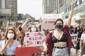 A group of people is gathered in a public area, holding various signs with messages advocating for women's rights and against assault. Many are wearing face masks, and there is a mix of emotions displayed, including determination and solidarity. The backdrop includes urban buildings with several people raising their fists in the air.