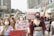A group of people is gathered in a public area, holding various signs with messages advocating for women's rights and against assault. Many are wearing face masks, and there is a mix of emotions displayed, including determination and solidarity. The backdrop includes urban buildings with several people raising their fists in the air.