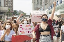 A group of people is gathered in a public area, holding various signs with messages advocating for women's rights and against assault. Many are wearing face masks, and there is a mix of emotions displayed, including determination and solidarity. The backdrop includes urban buildings with several people raising their fists in the air.