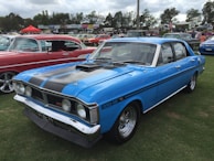 A classic blue car is prominently displayed on a grassy field, surrounded by other vintage vehicles. The car has a shiny exterior with black racing stripes and chrome details. In the background, several people are gathered, possibly at an outdoor car show event, with various tents and signs visible.