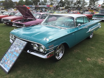 A teal vintage car is showcased on a lawn with a display of photographs leaning against the front. Other vintage cars are parked nearby, some with their hoods open. The setting resembles a car show with several people in the background.