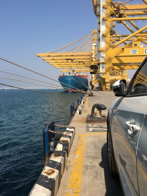 A large cargo ship is docked at an industrial port with massive yellow cranes overhead. The water is calm and blue, with a clear sky above. A white car is parked close to the edge of the quay, highlighting the industrial nature of the scene.
